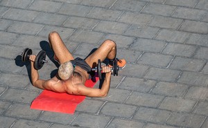 A shirtless man laying on a yoga mat and lifting weights.