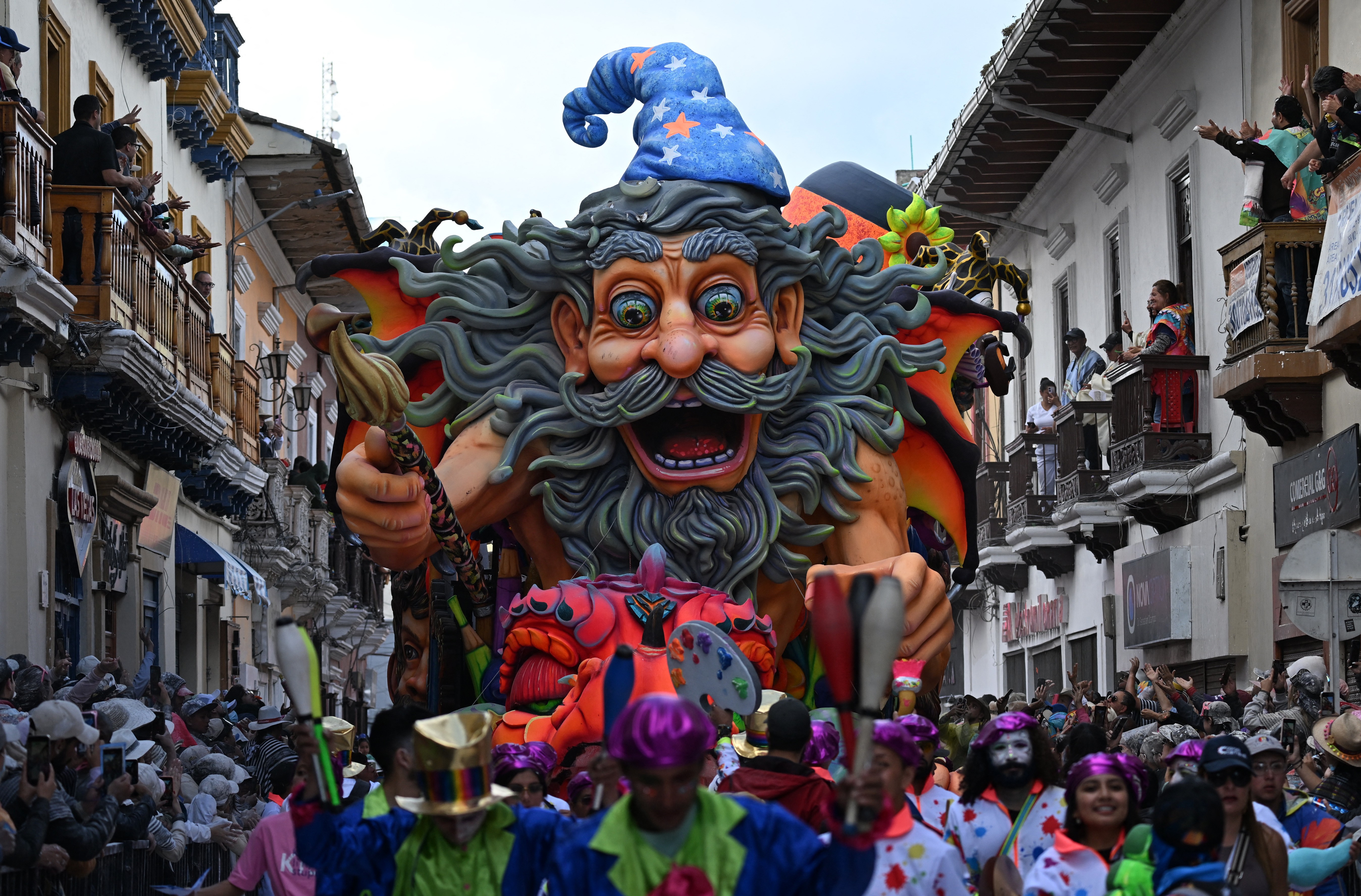 Costumed people parade in front of a large colorful float in the shape of a wizard.