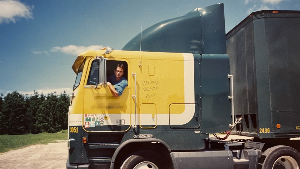 A man leans out of the window of a large truck with a blue and yellow cab.