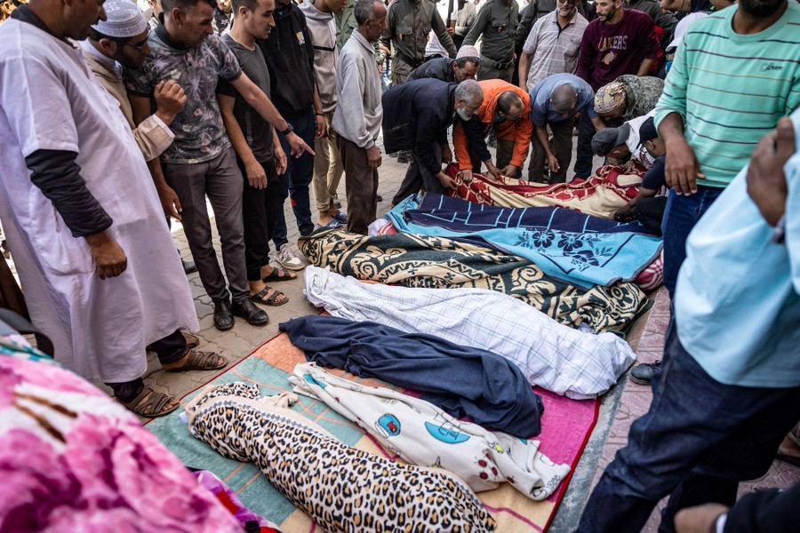 A crowd gathers around at least seven bodies, fully wrapped in blankets, laid on the ground.