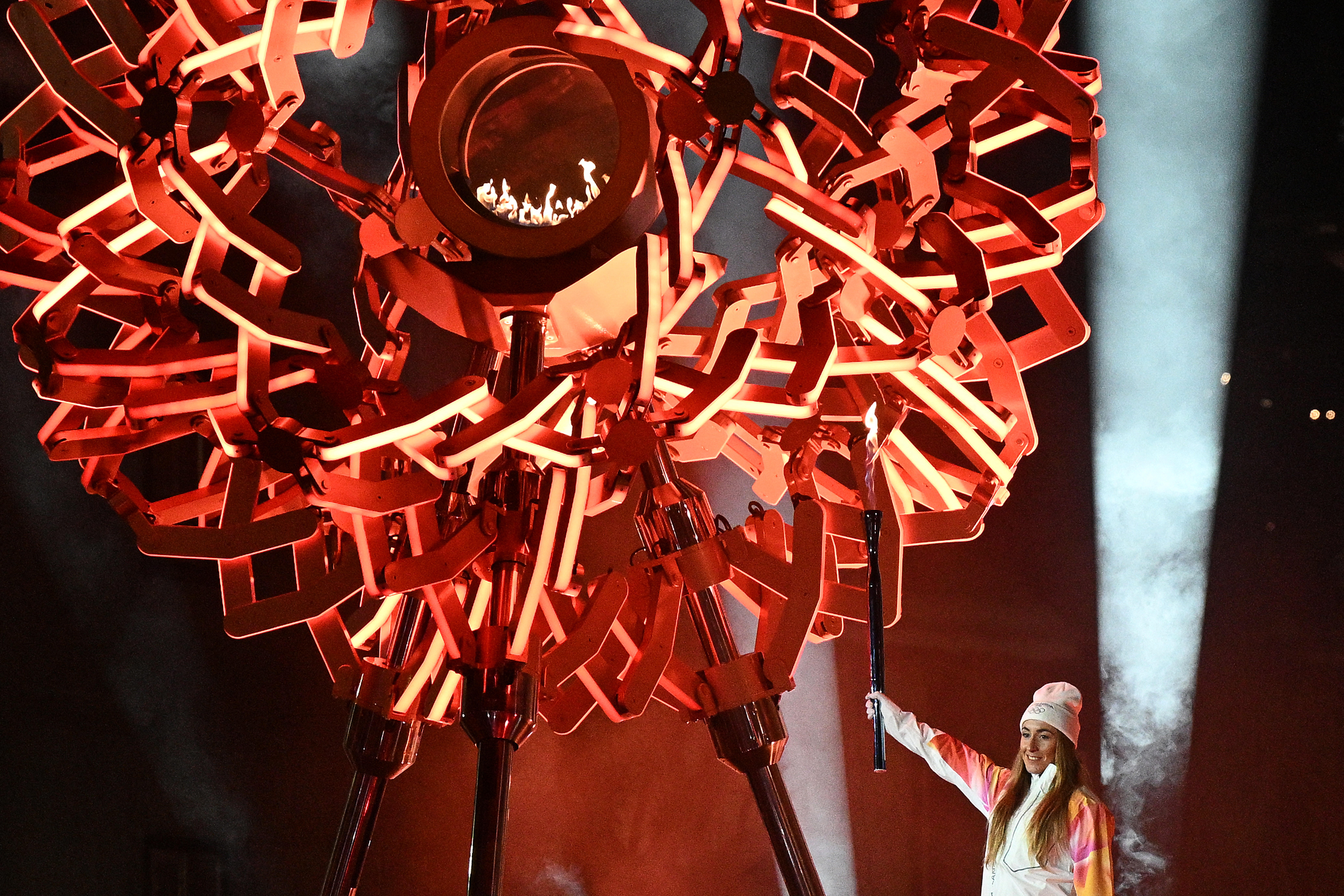 A torchbearer reaches up to ignite a large, ornate cauldron.