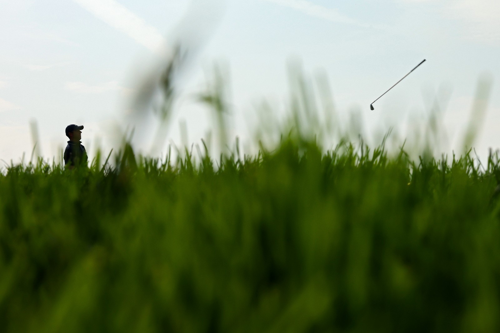 Golfer Rory McIlroy throws a club, seen through grass at a distance.