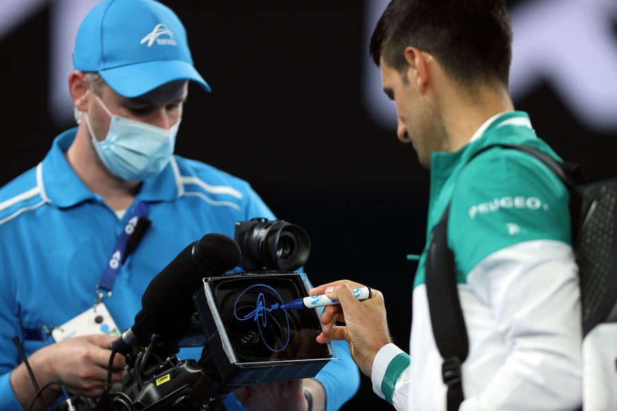 Novak Djokovic signs a piece of glass over the lens of a video camera being held by a camera operator.