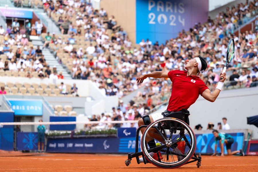 A para-athlete in a wheelchair reaches back to serve during a tennis match.