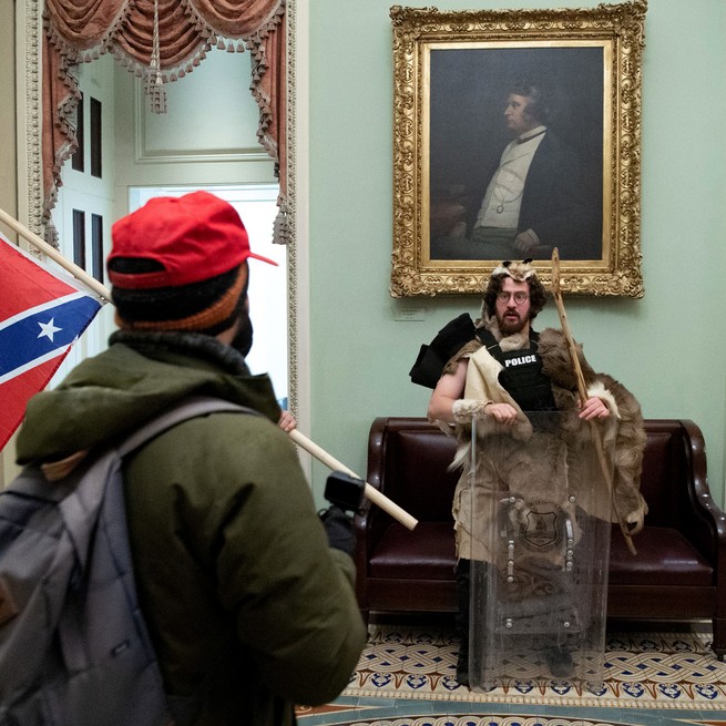 man wearing pelts stands in capitol building while a man carrying confederate flag looks on.