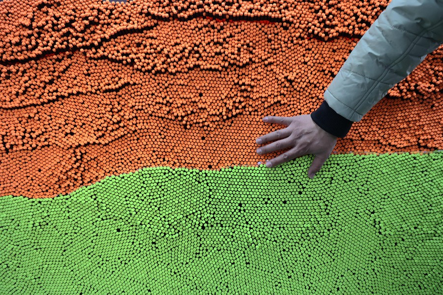 A person places their hand on a huge stack of orange and green colored pencils in a factory.