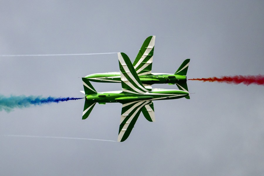 Two green-and-white striped military jets fly closely past each other, trailing colored smoke during an air show.