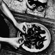 A black and white photo of someone cleaning mussels, with someone else's shoes in the photo, in black strappy sandals