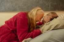 photo of older woman with long hair sitting on edge of bed and hugging man lying propped up on pillows