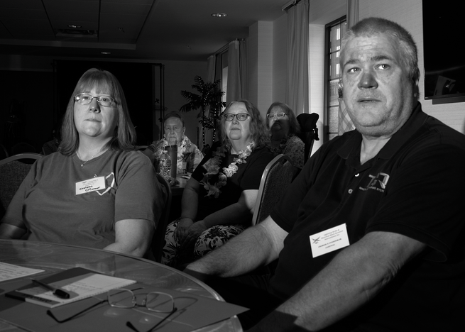 black-and-white photo of woman with glasses and man in dark t-shirt, both wearing conference badges, seated at table with more attendees wearing leis in background