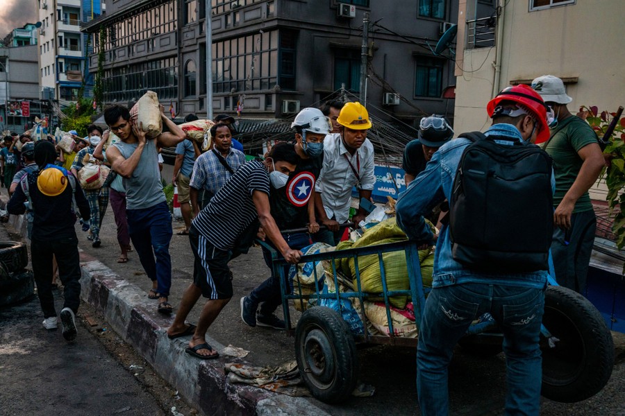 Several people carry and push carts full of sandbags on a city sidewalk.