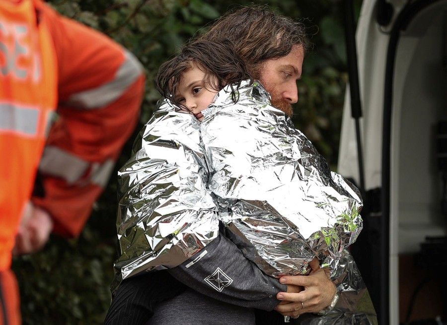 A father carries his young child, both wrapped in Mylar emergency blankets.