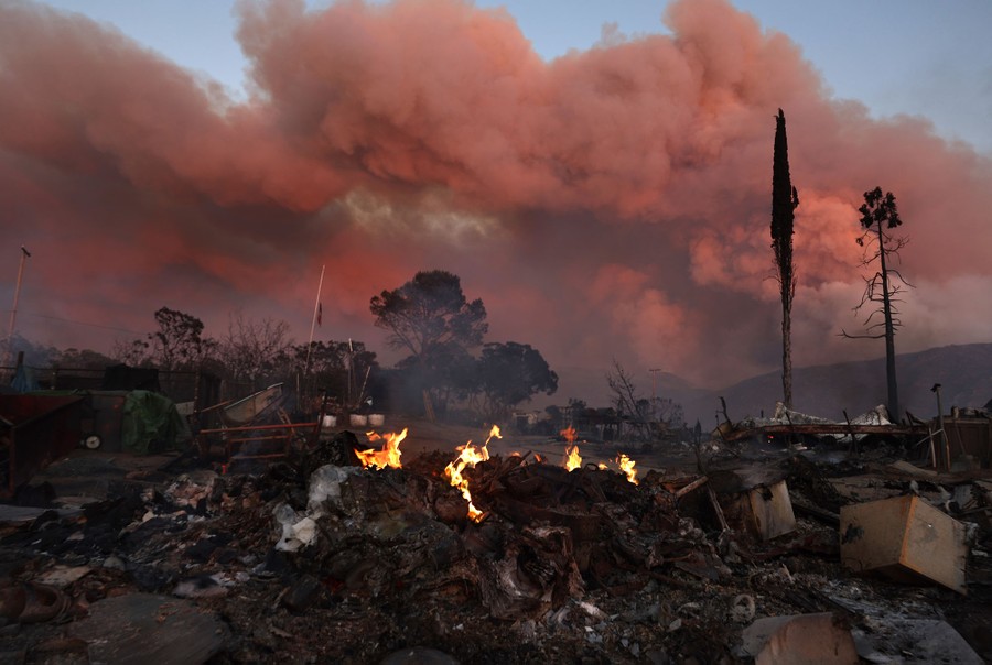 The burning and smoldering ruins of a building, against the backdrop of a large plume of smoke from a wildfire