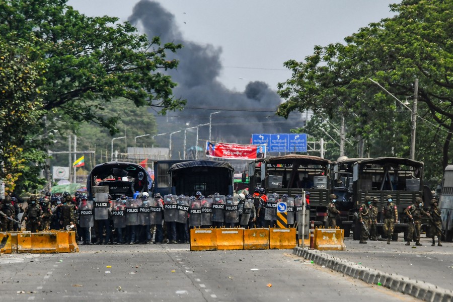 A large group of police officers stand together in a street, near transport trucks and barricades, with dark smoke in the background.
