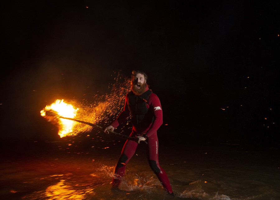 A bearded man in a wetsuit waves a torch as he stands in shallow water at night.