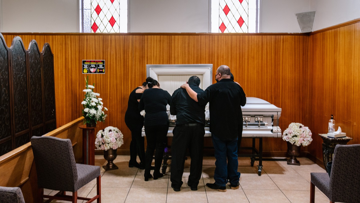 Family members dressed in black stand around a white casket, mourning at a funeral home.