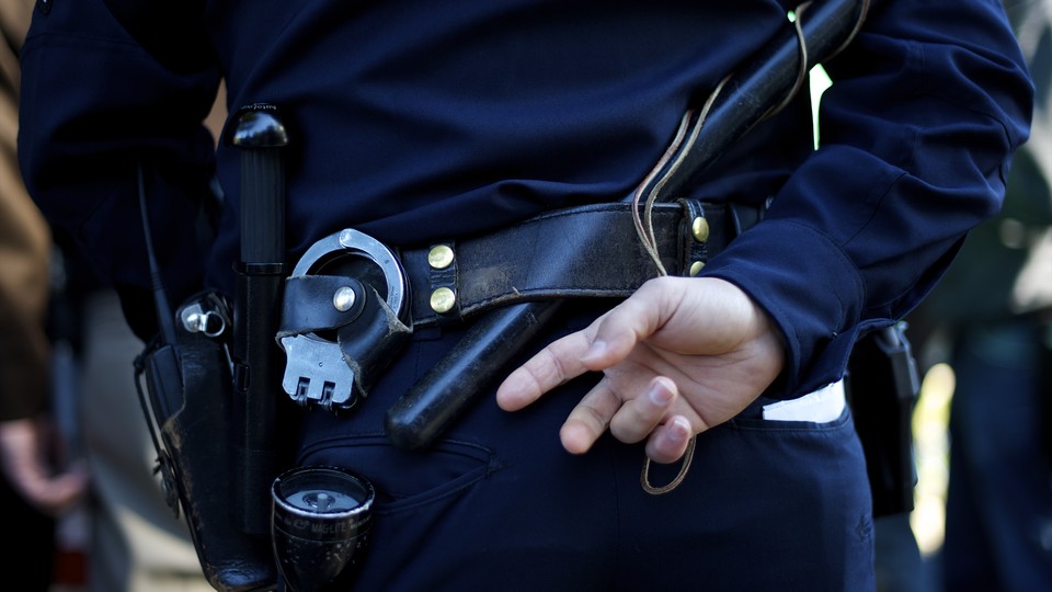 A police officer stands with one hand behind his back, with his handcuffs, flashlight, and radio visible.