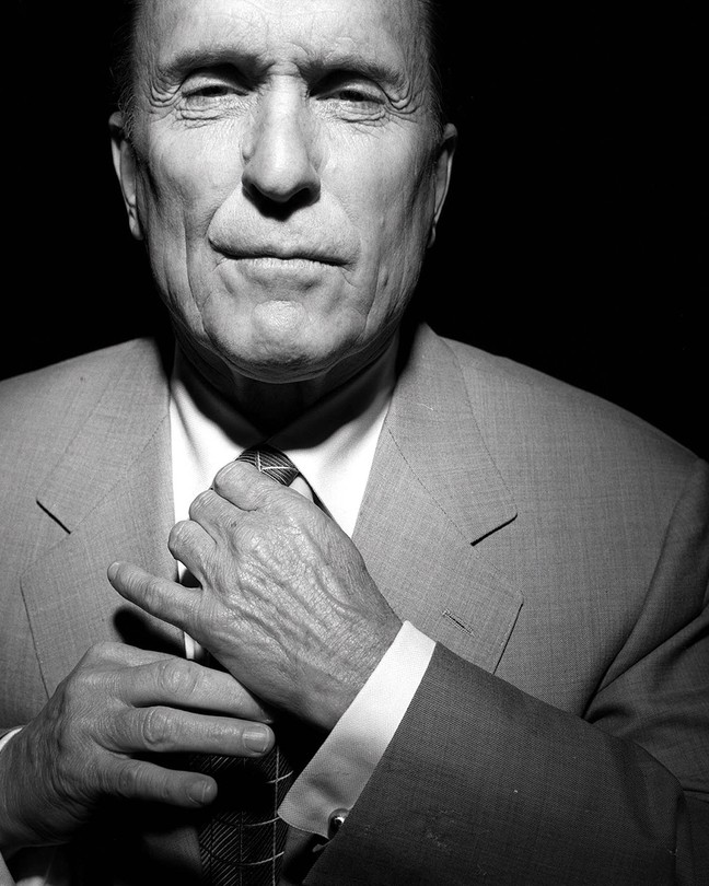 A black-and-white photograph of Robert Duvall wearing a suit and adjusting his tie.