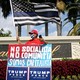 A man in a MAGA hat stands with a sign that reads "Not socialist, not communist, we are capitalist" in Spanish