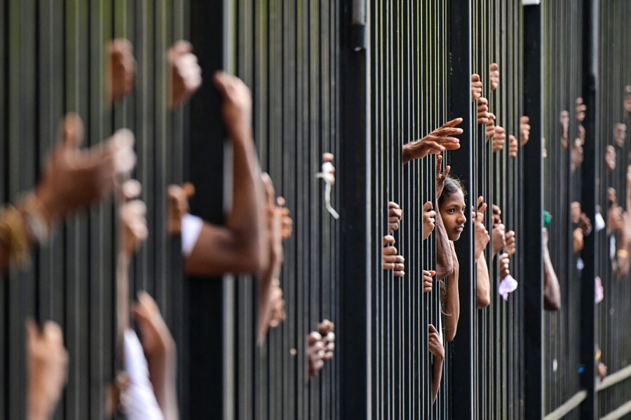 The hands and faces of many people are seen through the bars of a tall fence.