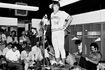 A black-and-white photo of Pete Rose addressing reporters in a locker room