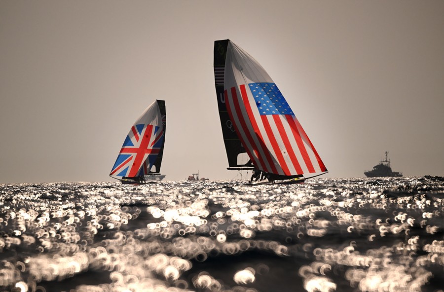 Two racing skiffs with sails adorned with country flags, in a distant view across water, with many reflections