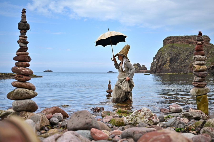A woman in costume wades near a rocky shore where stones are balanced in small towers.