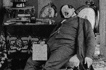 Old, black and white photograph of a man asleep in a chair with his mouth wide open.