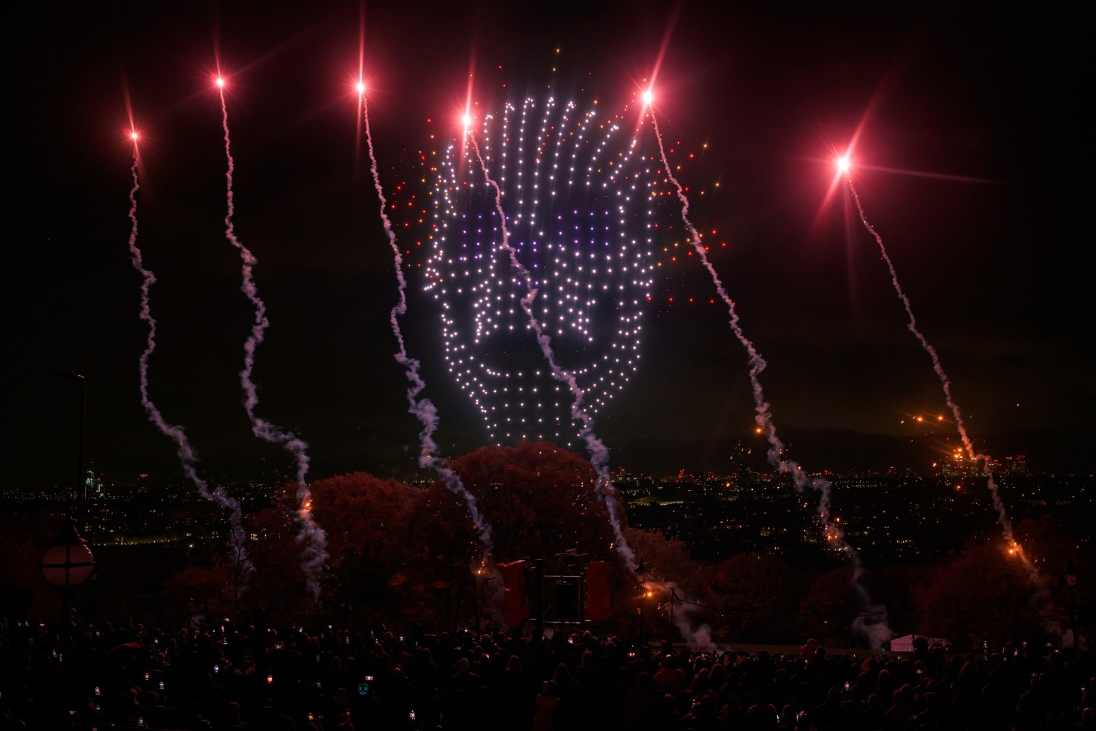 Fireworks erupt as a drone light show creates the shape of a human skull above a crowd.