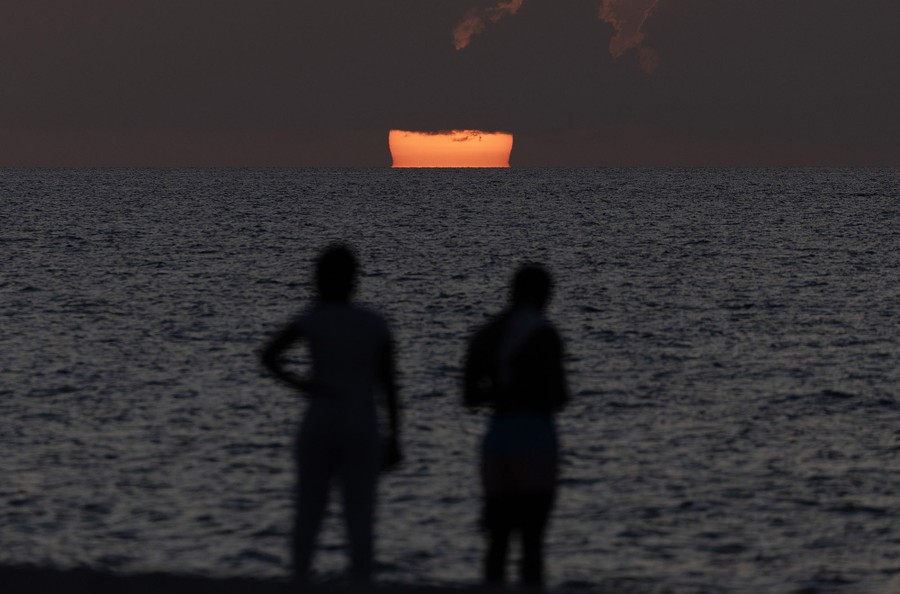 Two people are seen in silhouette watching the sun rise over the ocean.