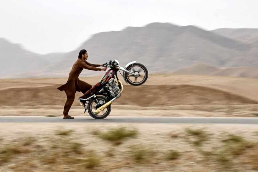 A man wearing sandals performs a trick on a motorcycle, popping its front wheel up and sliding along on one foot.