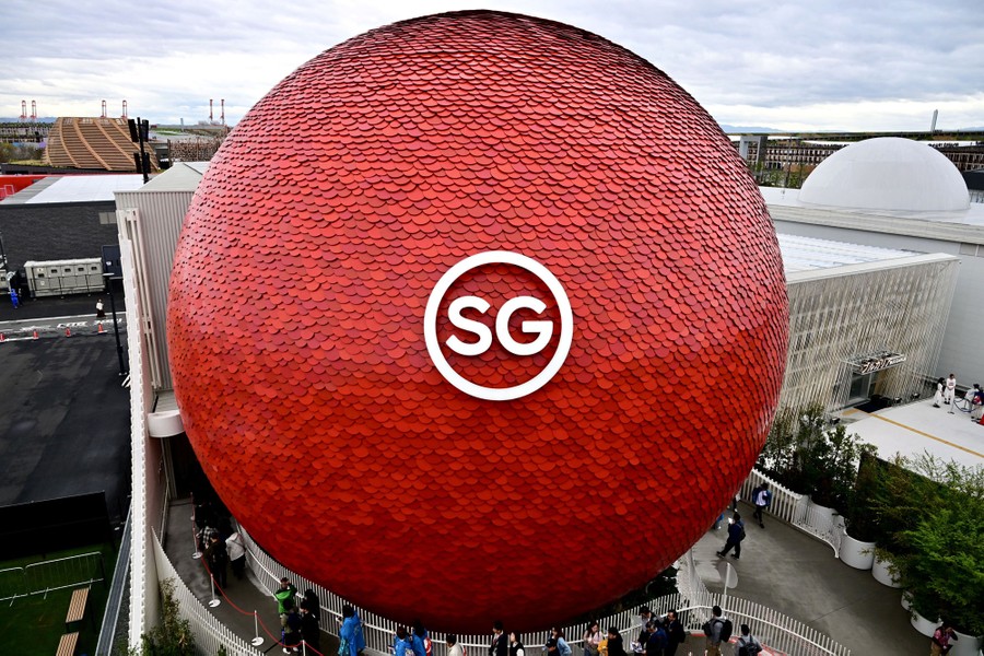 People walk beside a giant spherical structure covered by thousands of bright red tiles, bearing a large sign reading "SG."