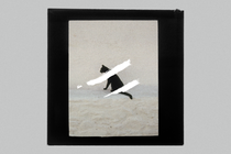 a cat in the distance, sitting on the sand, seen in profile. the image of the cat is covered in two white smudges of paint, and framed by a black square on a grey background
