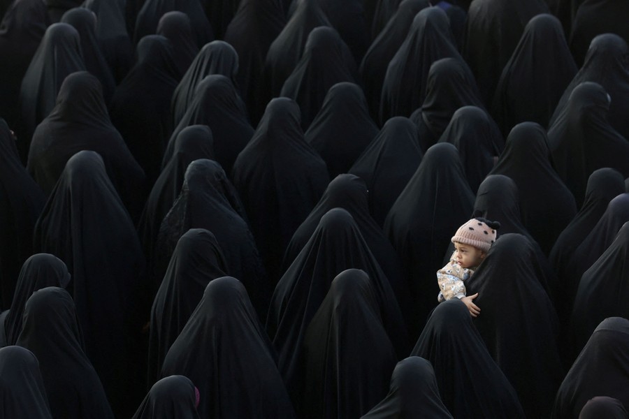 A crowd of women wearing black hijabs stand together. One holds a small child.