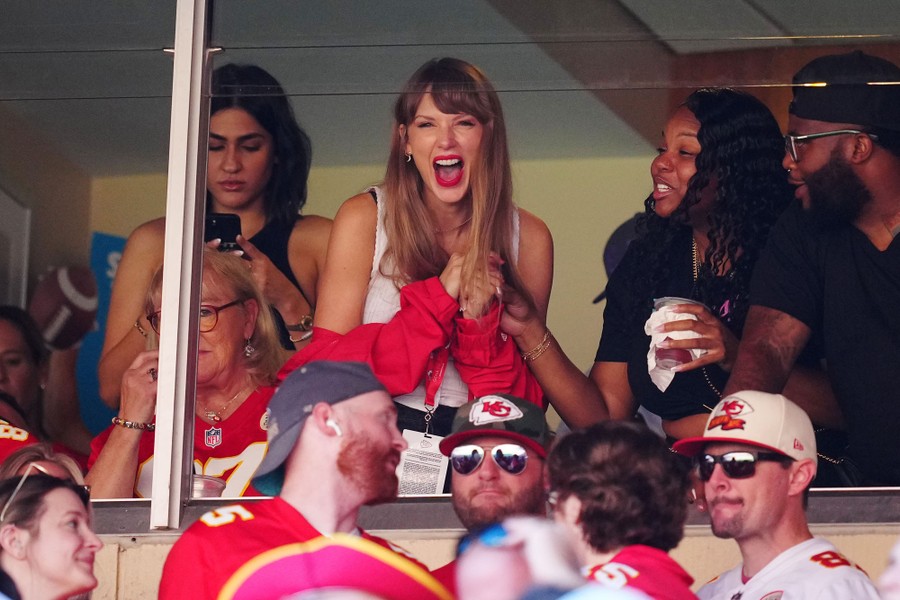 Taylor Swift and several others react to a football game, standing in a booth.
