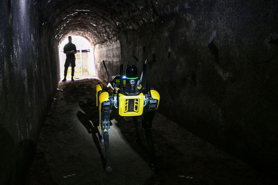 A person stands at the entrance to a tunnel, using a remote control to drive a four-legged robot into the tunnel.