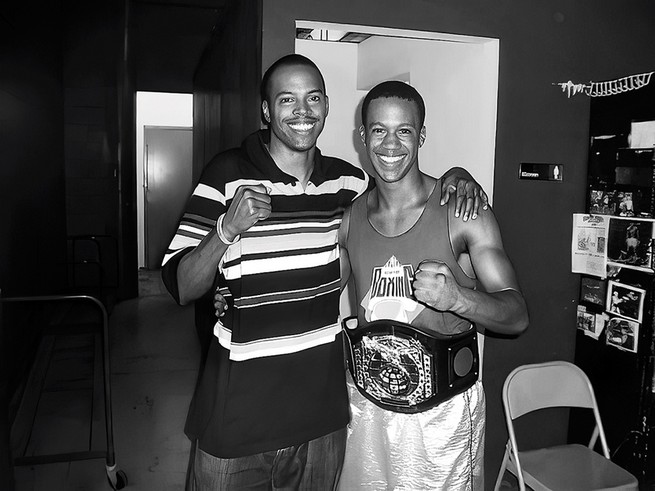 photo of smiling man in striped t-shirt with right hand in a fist and left arm around shoulder of man in boxing gear, also smiling with fist raised and wearing title belt