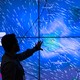 A meteorologist points to a big blue screen displaying wind data from hundreds of weather stations