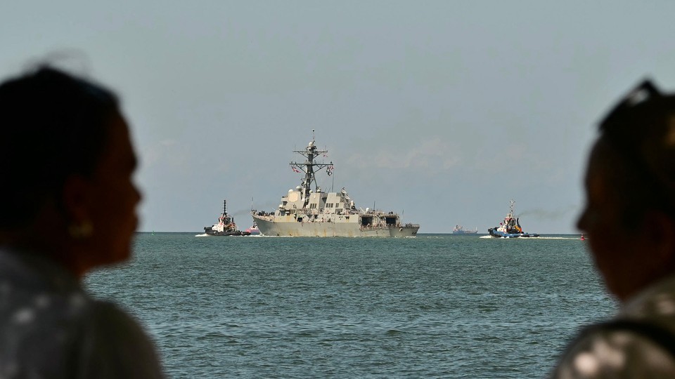Two people in the foreground look out at a U.S. Navy warship in the water at a distance.