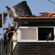 A color photograph of a man peering out the window of a house whose roof appears to have been destroyed by an explosion.
