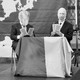 Two men play cards on a stage in front of a map of Europe, their faces obscured by cut-outs of Trump and Putin.