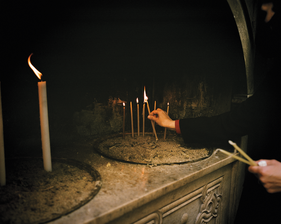 photo of hand reaching in from side and lighting thin candle by touching a lit candle, with a much larger candle burning nearby