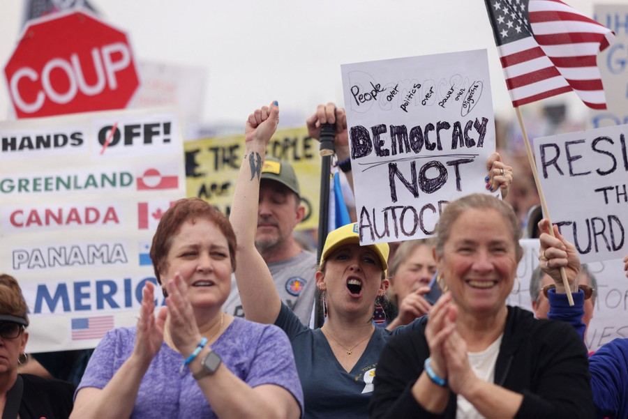 Protesters cheer and hold up signs, including one that reads "Democracy not autocracy."