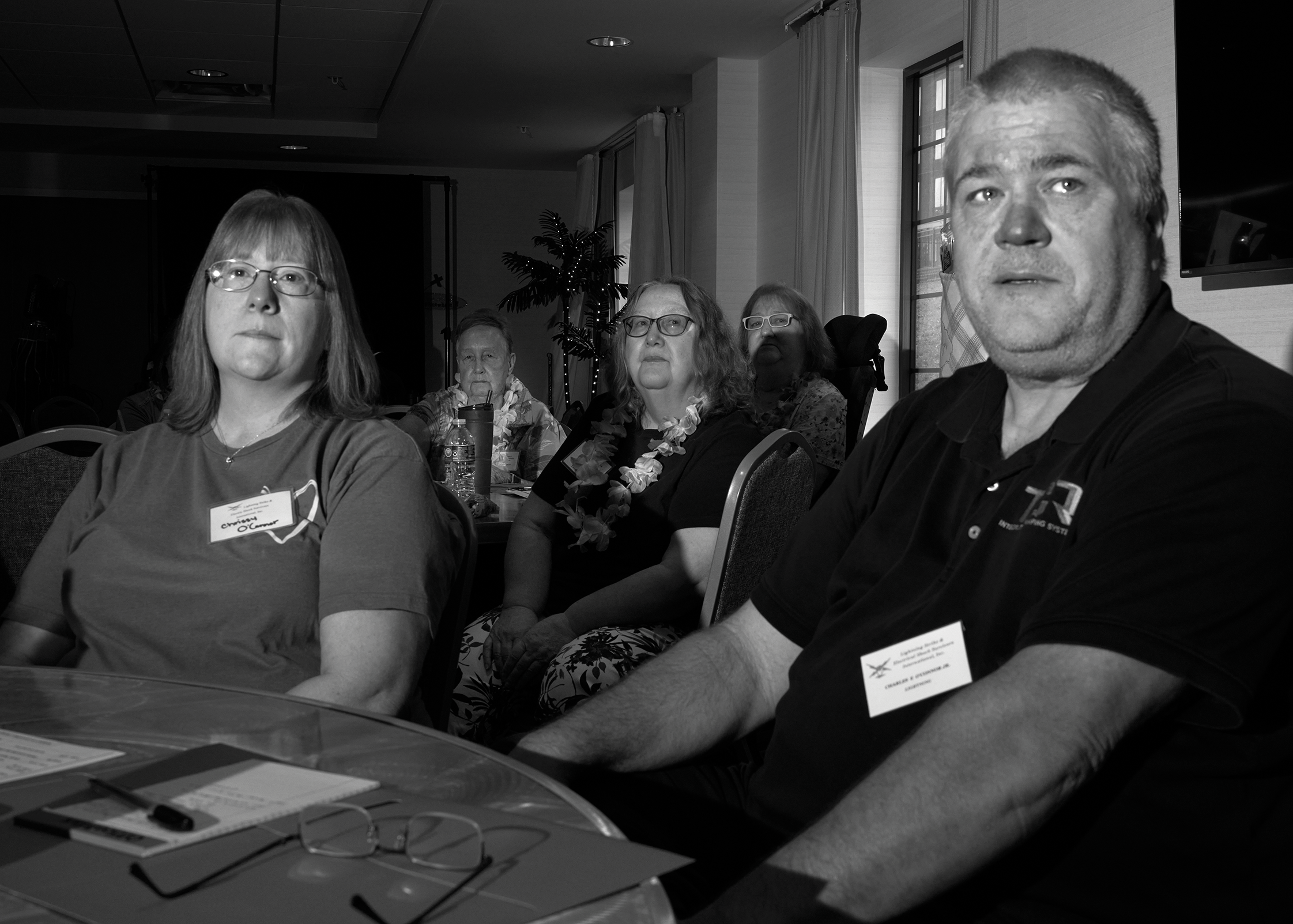 black-and-white photo of woman with glasses and man in dark t-shirt, both wearing conference badges, seated at table with more attendees wearing leis in background