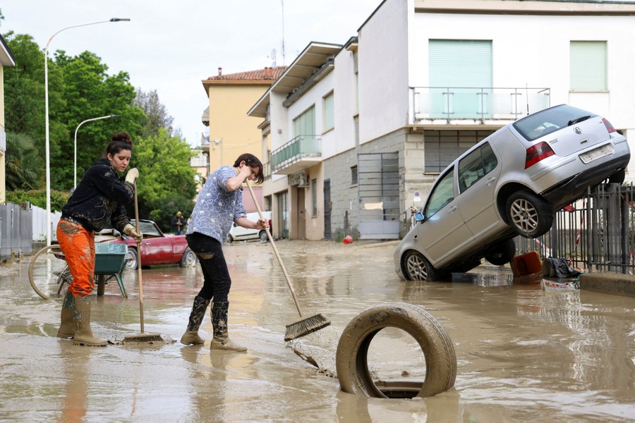 Two people sweep mud toward a drain as flood-damaged cars are perched nearby.