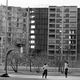A black and white photograph of two figures playing basketball in front of a ruined housing project