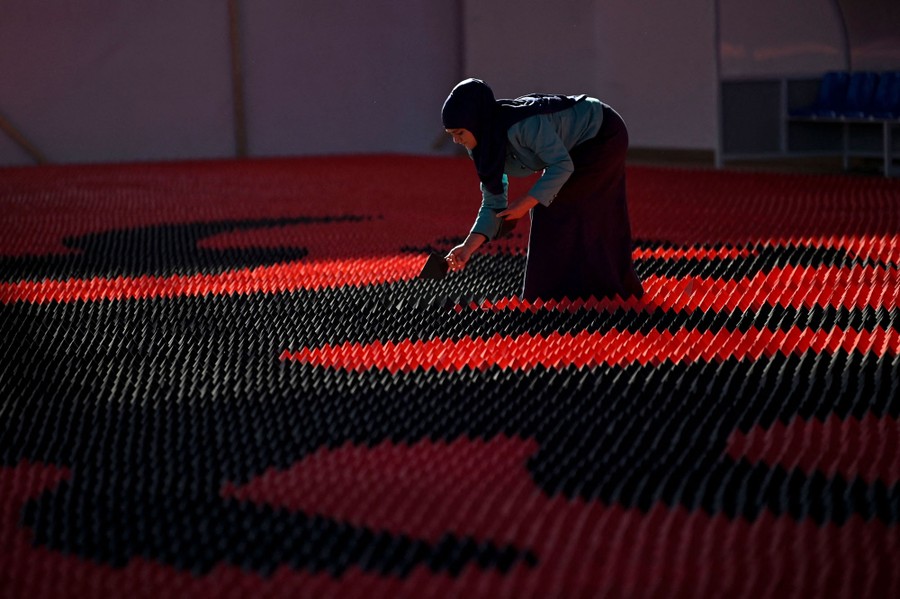 A person bends over inside a large room, gently placing small bits of folded paper on the floor, making a giant mosaic