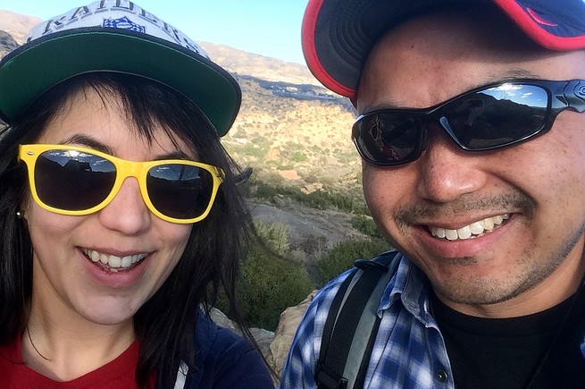 A woman and a man wearing baseball caps and sunglasses, close up and smiling in front of a mountain landscape