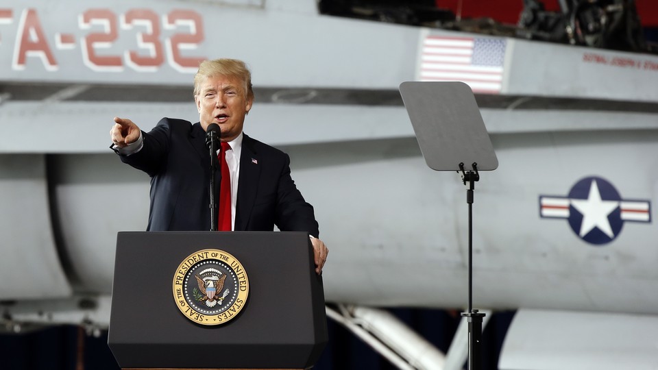 Trump stands at a podium and points while in front of an fighter jet.