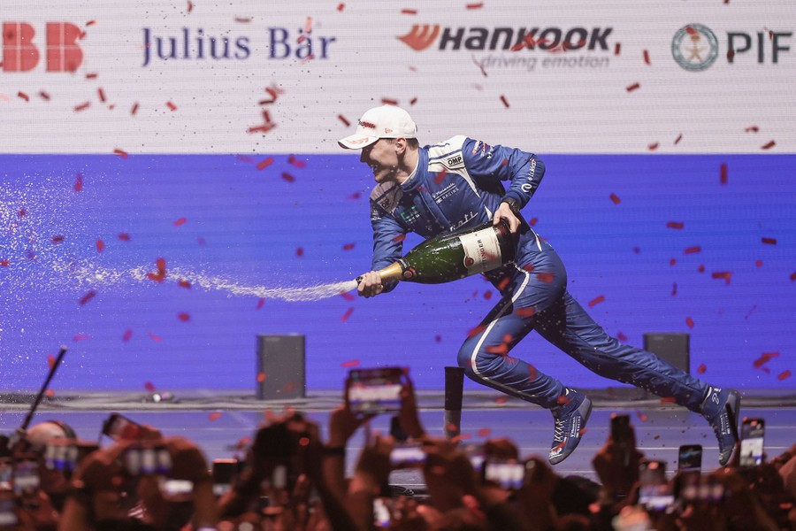 A race-car driver runs on a stage, celebrating, while spraying champagne from a huge bottle.